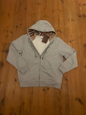 Grå Burberry Zip-Hoodie - Uppgradera din garderob med denna exklusiva Burberry zip-hoodie i en stilren grå nyans. Tillverkad med högkvalitativa material och Burberrys klassiska känsla för detalj och komfort — perfekt för både vardag och trendiga outfits. Bättre pris vid snabb affär!