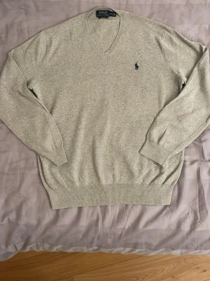 Ljusgrå stickad tröja Polo Ralph Lauren - Ljusgrå stickad tröja från Polo Ralph Lauren med klassisk v-ringning och broderad marinblå logga på bröstet. Tröjan har långa ärmar och ribbade muddar vid ärmslut och nederkant. Perfekt för en stilren och avslappnad look.