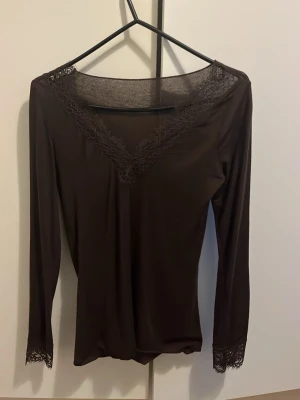 Intimissimi topp brun - Mörkbrun långärmad topp med v-ringning och fina spetsdetaljer vid halsringningen och ärmsluten. Tunn och stretchig modell som sitter snyggt på kroppen. Perfekt att styla med jeans eller kjol.