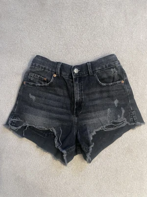 Svarta denimshorts med slitningar - Snygga svarta jeansshorts med råa kanter och slitna detaljer framtill. Klassisk femficksmodell med knapp och dragkedja. Shortsen har en något högre midja och är perfekta för en avslappnad stil.