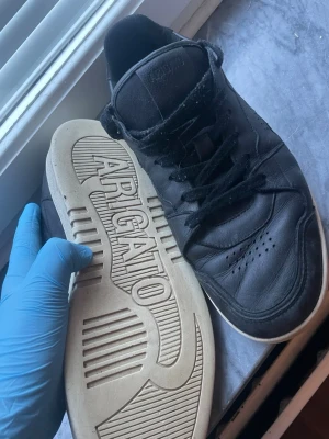 Svarta sneakers från Axel Arigato - Snygga svarta sneakers från Axel Arigato med klassisk låg profil och vit sula. Skorna är gjorda i slätt läder med perforerade detaljer på tån och har svarta skosnören. Märkets logga syns tydligt på sulan och sidan.