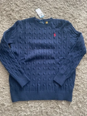 Blå kabelstickad tröja Polo Ralph Lauren - Snygg blå kabelstickad tröja från Polo Ralph Lauren med rund halsringning och den klassiska röda logotypen broderad på bröstet. Tröjan har långa ärmar och ribbade muddar vid ärmslut och nederkant. Perfekt för en stilren och avslappnad look.