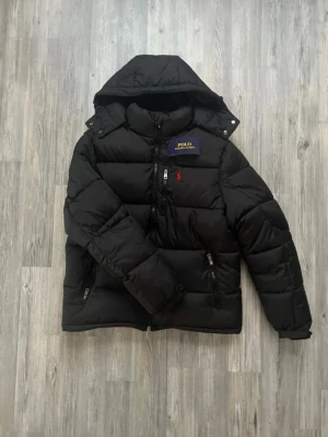 Svart dunjacka från Polo Ralph Lauren - Helt ny utan prislapp, Svart dunjacka från Polo Ralph Lauren med huva och dragkedja framtill. Jackan har två sidofickor med dragkedja och en bröstficka. Klassisk pufferstil med diskret röd logga på bröstet och quiltad design. Perfekt för kalla dagar.