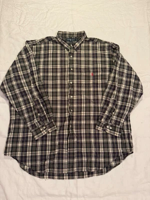 Rutig skjorta från Ralph Lauren - Klassisk rutig skjorta från Ralph Lauren i mörka toner med inslag av vitt, grönt och rött. Skjortan har knappar framtill, lång ärm och den ikoniska röda loggan broderad på bröstet. Tillverkad i 100% bomull och har en avslappnad passform.