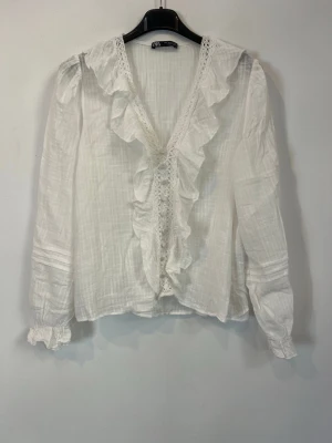 Vit blus med volanger och spets - Söt vit blus med djup V-ringning, volangdetaljer och spets längs kanten. Tillverkad i ett tunt, lätt material som ger en luftig känsla. Långa ärmar med resår vid ärmslut för extra stil. Perfekt för dig som gillar spets och volang detaljer.