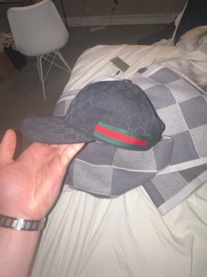 Svart Gucci keps med GG-mönster - Snygg svart keps från Gucci med klassiskt GG-monogram över hela kepsen och en grön-röd rand på sidan. Justerbar rem bak och insida i svart tyg. Perfekt för dig som vill ha en exklusiv och trendig accessoar.