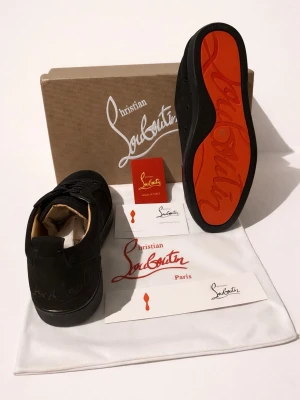Christian Louboutin skor Helt Nya!!! - Säljer ett par svarta sneakers från Christian Louboutin med ikonisk röd sula. Skorna har rund tå, snörning och är tillverkade i skinn med mattsvart finish. Kommer med originalkartong, dustbag och äkthetskort. Perfekta för dig som vill ha något extra till din outfit.                                                                         Helt Nya!!!