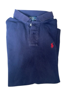 Marinblå pikétröja från Polo Ralph Lauren - Klassisk marinblå pikétröja från Polo Ralph Lauren med röd broderad logga på bröstet. Tröjan har korta ärmar, krage och knäppning med två knappar framtill. Tillverkad i bomull för en bekväm och stilren look.