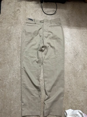 Beige kostymbyxor med raka ben - Snygga beige kostymbyxor med klassisk rak passform och bälteshällor. Byxorna har diskreta fickor fram och bak, samt stängs med knapp och dragkedja. Perfekta för en clean och stilren look.