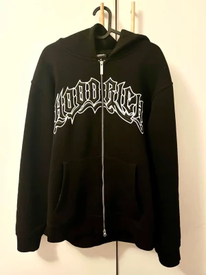 Svart zip hoodie från Hoodrich - Svart hoodie med dragkedja från Hoodrich. Stor vit broderad logga i streetstil över bröstet. Klassisk huva och stora fickor framtill. Tillverkad i mjukt bomullsmaterial som känns skönt mot huden. 