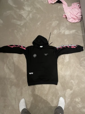 Off White hoodie  - Svart hoodie från Off-White med huva och stora rosa tryck på både ärmar och rygg. Textdetaljer i vitt och rosa, samt ikoniskt Off-White-motiv på ryggen. Hoodien har lång ärm och klassisk passform. Perfekt för dig som gillar streetwear med attityd. Stl L passar M