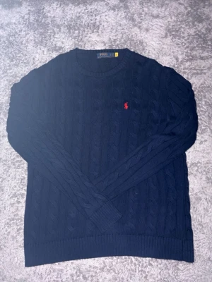 Mörkblå kabelstickad tröja Polo Ralph Lauren - Mörkblå kabelstickad tröja från Polo Ralph Lauren med rund halsringning och röd broderad logga på bröstet. Klassisk modell med ribbade muddar och nederkant. Perfekt för en stilren och avslappnad look. 