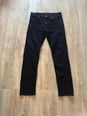 Svarta raka jeans från Lee - Säljer ett par svarta jeans från Lee med rak passform och klassisk femficksdesign. Jeansen har knappgylf och Lee-logga bak i midjan. Tillverkade i ett mjukt och slitstarkt denimtyg som passar perfekt till en avslappnad stil. Storlekarna är W33 och L34 vilket passar mig som brukar ha storlek L i byxor.