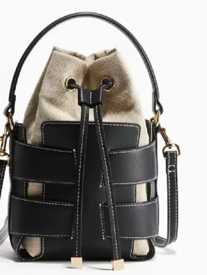 Beige och svart bucket bag med snörning - Trendig bucket bag i beige canvas och svart skinn med vita sömmar. Väskan har snörning upptill, guldfärgade detaljer och både handtag och axelrem. Perfekt för dig som vill ha en cool och stilren accessoar.