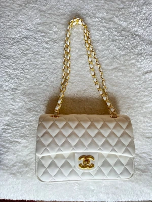 Vit quiltad axelväska från Chanel - Supersnygg vit axelväska från Chanel med klassiskt quiltat mönster och guldfärgad kedja. Väskan har ikonisk CC-detalj framtill och är tillverkad i skinn. Perfekt för dig som vill ha en tidlös och lyxig accessoar.