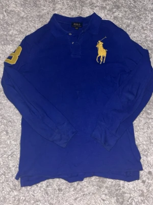 Blå långärmad Polo Ralph Lauren henley - Klassisk blå långärmad henley från Polo Ralph Lauren med stor gul logga på bröstet och en gul siffra på ärmen. Skjortan har krage och knappar upptill. Perfekt för en avslappnad men ändå stilren look.
