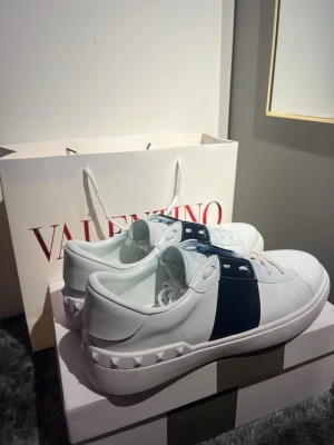Valentino Garavani Open sneakers vit/blå - Säljer ett par Valentino Garavani Open sneakers i vitt skinn med bred marinblå rand över vristen. Klassisk låg modell med vita snören och coola nitar på hälen. Perfekta för dig som vill ha en clean men ändå statement look. !OBS! Inte orginal snören då dom gick sönder. Men skon är i nyskick