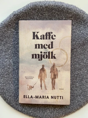 Kaffe med mjölk av Ella-Maria Nutti - Boken är ny och oläst! En roman om relationen mellan en mamma och en dotter, med teman kring avstånd, familjeband och hemligheter. Omslaget är i mjuka beige och grå toner med silhuetter och kaffefläckar (kaffefläckarna hör omslaget till!) Perfekt för dig som gillar starka berättelser om familj.