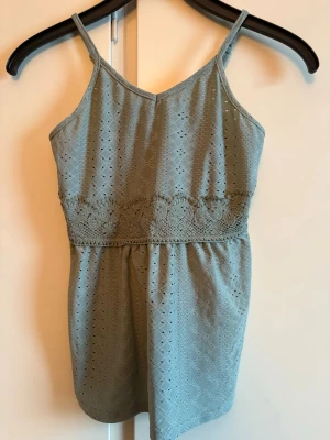 Grön spetsdetaljerad linnetopp - Snygg ljusgrönt linne med tunna axelband som man kan justera och broderade spetsdetaljer under bysten. Är hålmönster över hela tyget och är lätt utsvängd nedtill. Aldrig använt 