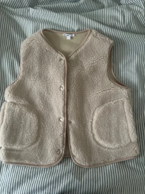 Beige teddyväst från Subdued - Mysig beige teddyväst från Subdued med två stora fickor framtill och tryckknappar. Västen har en rund halsringning och är tillverkad i ett mjukt, fluffigt material som ger en trendig look. Perfekt att slänga över en hoodie eller långärmad tröja. Aldrig använd 