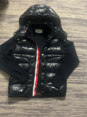 Svart dunväst med huva från Moncler - Snygg svart dunväst från Moncler med glansig finish, avtagbar huva och stickade ärmar. Dragkedjan har en röd, vit och blå rand som ger en sportig vibe. Perfekt för lager på lager och streetstyle. Klassisk Moncler-logga på insidan.