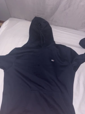 Svart hoodie från Tommy Hilfiger - Klassisk svart hoodie från Tommy Hilfiger med liten logga på bröstet. Hoodien har huva med dragsko och en stor magficka framtill. Perfekt för en avslappnad och stilren look.