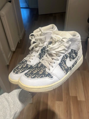 Nike Air Jordan 1 med mönstrad panel - Unika Nike Air Jordan 1 sneakers i vitt skinn med blått och beige mönster på sidor och tå. Klassisk snörning, rund tå och vit sula med gulnad kant för retrovibe. Swoosh-logga på sidan och högt skaft för ikonisk look.