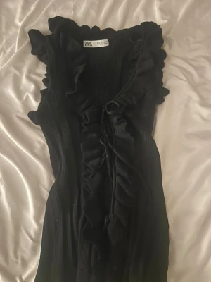 Svart volangprydd topp från Zara - Snygg svart topp från Zara med volangdetaljer längs urringningen och framtill. Toppen är ärmlös och har en ribbad struktur som ger en schysst passform. Perfekt för dig som vill ha något extra till outfiten.