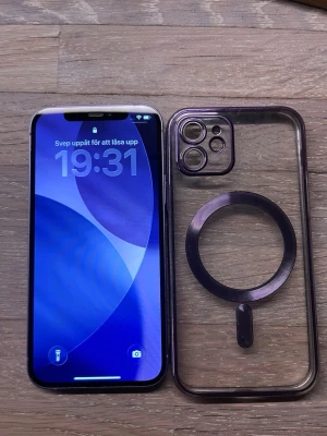 Lila iPhone 12 med MagSafe-skal - Säljer en snygg lila iPhone 12 med platt design och dubbla kameror. Baksidan är i glas och ramen i aluminium. Medföljer ett genomskinligt MagSafe-kompatibelt skal som har en lila ring för trådlös laddning. Perfekt för dig som vill ha en stilren och modern mobil.