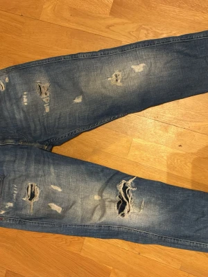 Blå slitna jeans med hål - Säljer ett par blå jeans med flera slitningar och hål på benen för en riktigt avslappnad och trendig look. Jeansen har en rak passform och klassisk femficksdesign. Perfekta för dig som gillar en edgy stil.