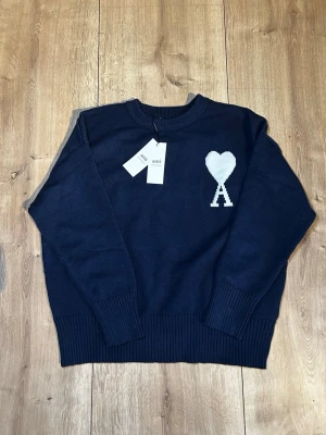 Mörkblå stickad tröja från Ami Paris - Snygg mörkblå stickad tröja från Ami Paris med deras ikoniska vita hjärta och A på bröstet. Tröjan har rund hals, ribbade muddar och nederkant. Perfekt för dig som gillar stilrena och trendiga plagg i garderoben.
