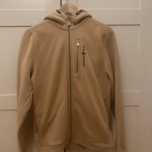 Beige hoodie från Sail Racing - Snygg beige hoodie från Sail Racing med dragkedja framtill och bröstficka med logga. Tröjan har huva med snörning och långa ärmar. Perfekt för dig som gillar stilren och sportig look.