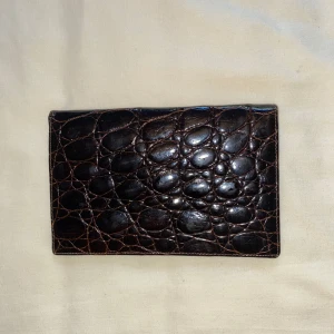 Brun plånbok i krokopräglat skinn - Snygg brun plånbok med krokopräglad yta i glansigt skinn. Plånboken har flera fack för kort och sedlar samt en smart knappstängning. Perfekt för dig som gillar klassisk stil med en twist. Passar enkelt i fickan eller väskan.