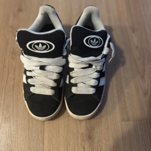 Adidas Campus svarta sneakers strl 40 - Svarta Adidas Campus sneakers med klassiska vita ränder och logga på plösen. Skorna har breda vita snören, gummisula i brunt och ovandel i mocka. Perfekta för dig som gillar retrostil och streetwear.