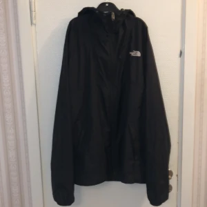 Svart vindjacka / regnjacka från The North Face - Svart vindjacka från The North Face med huva och vit broderad logga på bröstet. Jackan har dragkedja framtill och är tillverkad i lätt polyester, perfekt för blåsiga dagar. Stilren och enkel design med långa ärmar och normal passform.