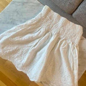 Vit broderad kjol från By Malina - Supersöt vit kortkjol från By Malina i storlek XS. Kjolen har bred smock i midjan och vackra broderade detaljer med spets längs nederkanten. Tillverkad i mjuk bomull. Perfekt för dig som gillar romantisk och feminin stil.