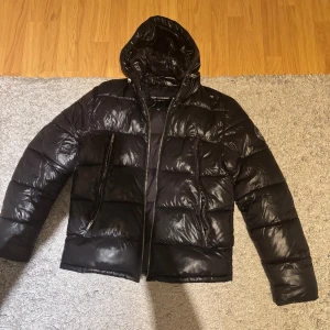 Svart pufferjacka Supply & Demand - Säljer en svart pufferjacka från Supply & Demand med glansig finish, huva och två stora dragkedjefickor framtill. Jackan har tydliga quiltade partier och en snygg logga på ärmen. Perfekt för kalla dagar när du vill hålla stilen.