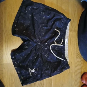 Louis Vuitton svarta shorts med snörning - Svarta shorts från Louis Vuitton med ikoniskt monogrammönster över hela tyget. De har elastisk midja med vit snörning och är tillverkade i ett lätt material som passar perfekt för sommaren. Stilen är loose och de har en sportig vibe.
