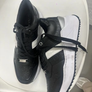 Svarta sneakers från Calvin Klein - Snygga svarta sneakers från Calvin Klein med präglad logga över hela skon. Skorna har vita och silvriga detaljer, svart snörning och en chunky vit sula. Tillverkade i skinnliknande material med modern och sportig vibe. Perfekta för dig som gillar stilrena och trendiga sneakers.