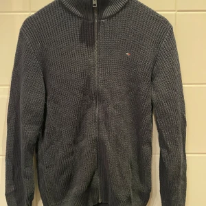 Mörkblå stickad kofta Tommy Hilfiger - Mörkblå stickad kofta från Tommy Hilfiger med dragkedja framtill och diskret logga på bröstet. Tröjan har ribbad krage och muddar samt långärmad passform. Perfekt för lager-på-lager under kyliga dagar.