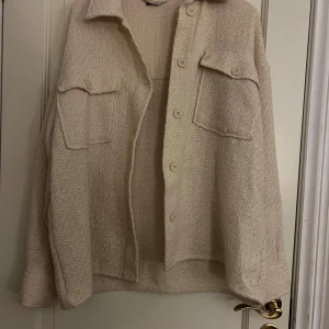 Beige overshirt med fickor och knappar - Stilren beige overshirt i grovstickat material med två stora bröstfickor och knäppning framtill. Jackan har en avslappnad passform och klassisk krage, perfekt för lager-på-lager. Materialet är strukturerat och mjuk. Det finns slitage på axel och  fläckar som man kan se på bilden.