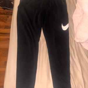 Svarta mjukisbyxor från Nike - Svarta mjukisbyxor från Nike med klassisk vit swoosh-logga på låret. Byxorna har en avslappnad passform och är tillverkade i mjukt material som känns skönt mot huden. Perfekta för chill eller träning. Står M men passar S mest 