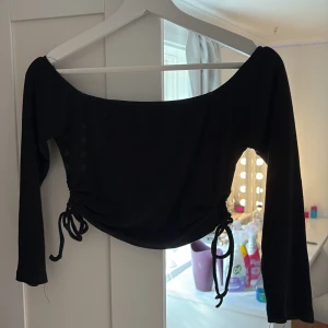 Svart offshoulder topp med snörning - Säljer en svart offshoulder topp med långa ärmar och trendig snörning i sidorna som ger en snygg rynkad effekt. Toppen är ribbad och har en croppad passform, perfekt att styla med höga jeans eller kjol. Materialet känns mjukt och stretchigt.