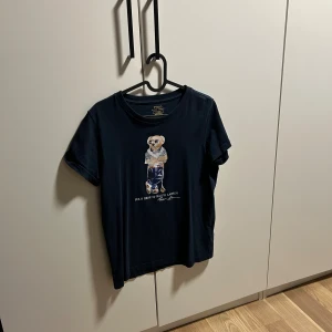 Mörkblå Polo Ralph Lauren t-shirt - Snygg mörkblå t-shirt från Polo Ralph Lauren med ikoniskt björntryck på bröstet. Klassisk rund hals och korta ärmar. Tillverkad i mjuk bomull som känns skön mot huden. Perfekt för dig som gillar streetstyle och vill sticka ut lite extra.