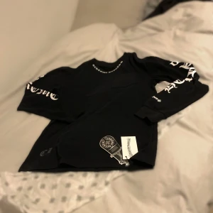 Svart långärmad tröja Chrome Hearts - Svart långärmad tröja från Chrome Hearts med vit logotyptext på ärmarna och runt halsen. Tröjan har en klassisk passform och är tillverkad i mjuk bomull. Perfekt för dig som gillar streetwear och vill sticka ut med snygga detaljer.