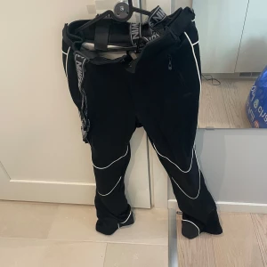 Svarta täckbyxor från Swix - Svarta täckbyxor från Swix med vita kontrastsömmar och dragkedja vid fickan. Byxorna har elastiskt midjeband med Swix-logga och är perfekta för kalla dagar. Snygg sportig look med detaljer längs benen. Använts som skidbyxor med tight passform