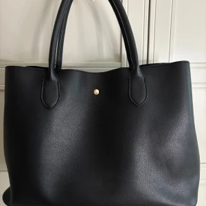 Svart axelväska i skinnimitation - Stilren svart axelväska i skinnimitation med två rejäla handtag och guldfärgade detaljer. Väskan har ett stort huvudfack, innerfack med dragkedja och tryckknappsstängning. Perfekt för dig som vill ha plats för allt du behöver under dagen.