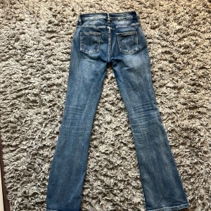 Blå lowaiste bootcut jeans - Snygga blå jeans med bootcut passform och klassisk femficksdesign. Jeansen har en något tvättad look och en trasig hällas bak i midjan. Perfekta för dig som gillar en avslappnad och trendig stil. Finns inga hål men den där grejen gick av lite