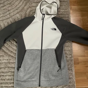 Grå och vit hoodiekofta från The North Face - Snygg hoodiejacka från The North Face i grått och vitt med svarta detaljer. Jackan har hel dragkedja, två fickor med dragkedja och en stor huva. Perfekt för dig som gillar sportig och clean stil.