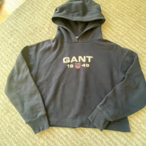 Svart hoodie från GANT med tryck - Svart hoodie från GANT med broderat GANT 1949-tryck och sköld på bröstet. Klassisk modell med huva, ribbade muddar och mjukt material. Perfekt för dig som gillar stilrena och sportiga plagg med premiumkänsla.